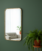 Rectangular mirror with gold frame on a green wall, reflecting a living room with a sofa and plants.