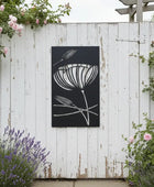 Decorative metal wall art with floral design on a white wooden fence surrounded by flowers.