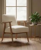 Beige armchair with wooden frame and legs, placed next to a small round table with a plant in a room with large windows.
