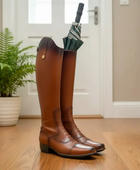 Brown riding boots with a green umbrella inside, placed on a wooden floor.