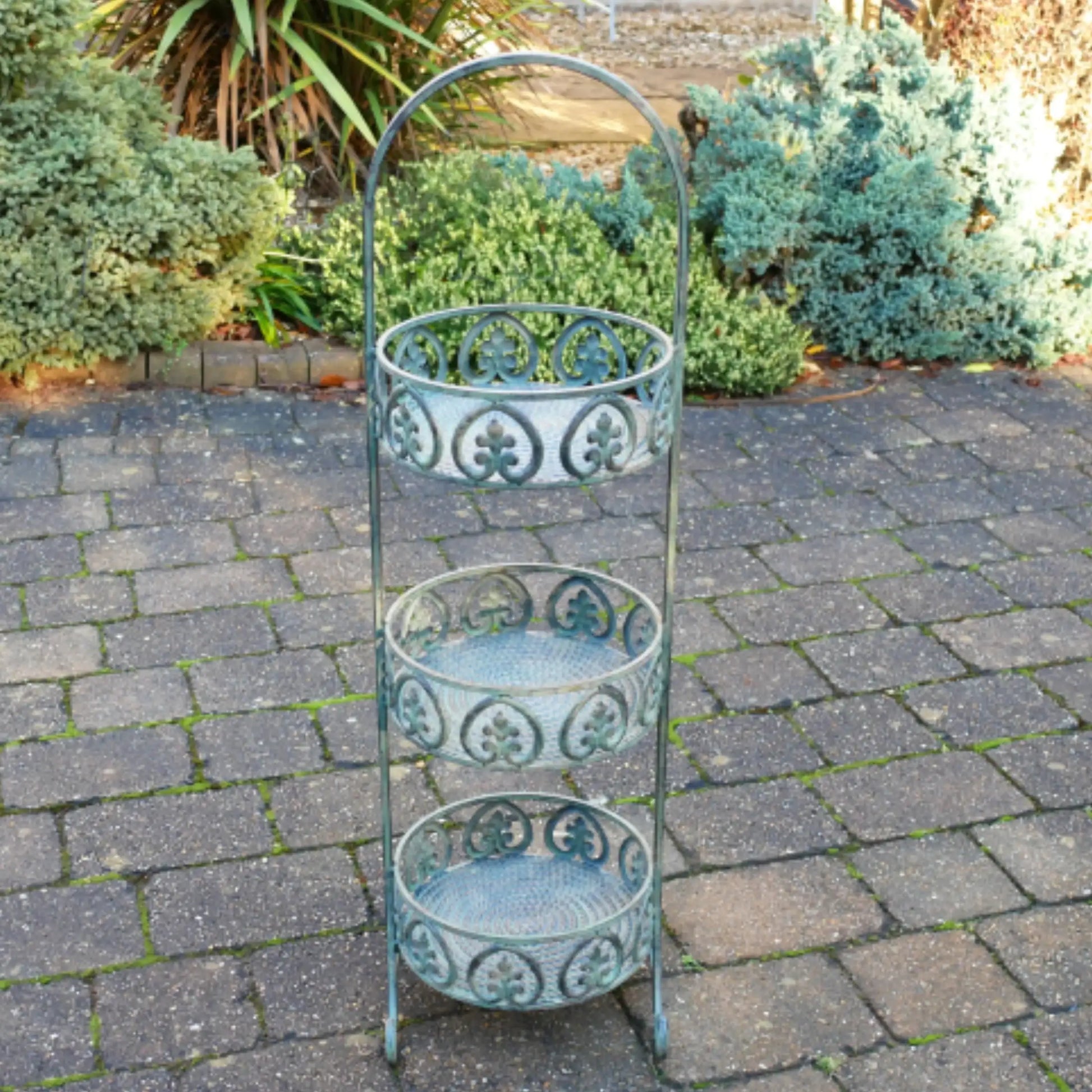 Decorative three-tier metal plant stand on a paved garden area with plants in the background.
