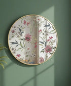 Round floral mirror with gold frame on a green wall next to a potted plant.