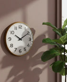 Round wall clock with gold frame on a beige wall next to a green plant