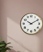Round wall clock with gold frame on a beige wall, next to a plant.