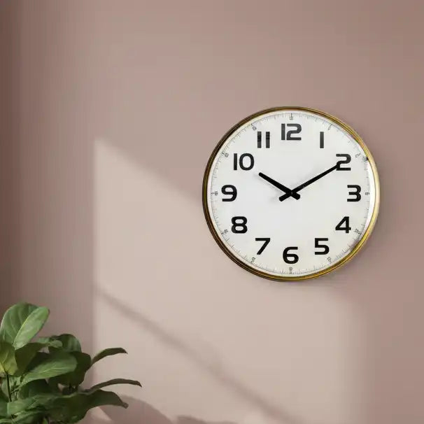Round wall clock with gold frame on a beige wall, next to a plant.