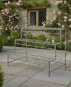 Decorative metal plant stand in a garden setting with flowers and a stone building.