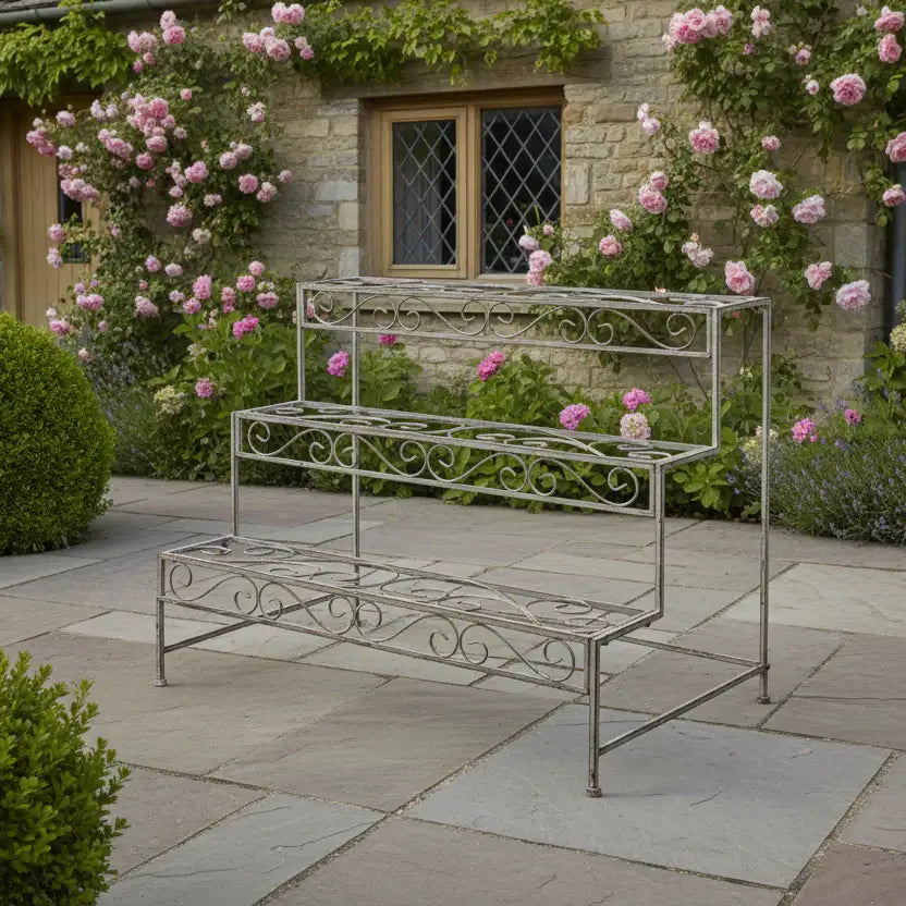 Wrought iron plant stand in a garden setting with pink flowers and a stone building.