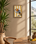 A framed art print depicting a yellow and white striped table setting with a plate of pancakes and a cup of coffee, hanging on a beige wall in a sunlit dining room next to a window and a potted plant.
