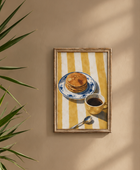 A framed art print featuring a breakfast scene with pancakes on a blue-and-white plate, a cup of coffee, and a spoon over a yellow-and-white striped background, hanging on a beige wall next to a green plant.