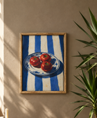 A framed art print depicting three red tomatoes on a blue and white striped plate, hung on a beige wall next to a green plant.
