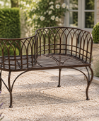 A dark brown wrought iron garden bench with a decorative lattice backrest, set on a gravel path in a landscaped garden with lavender, purple flowers, and green shrubs.