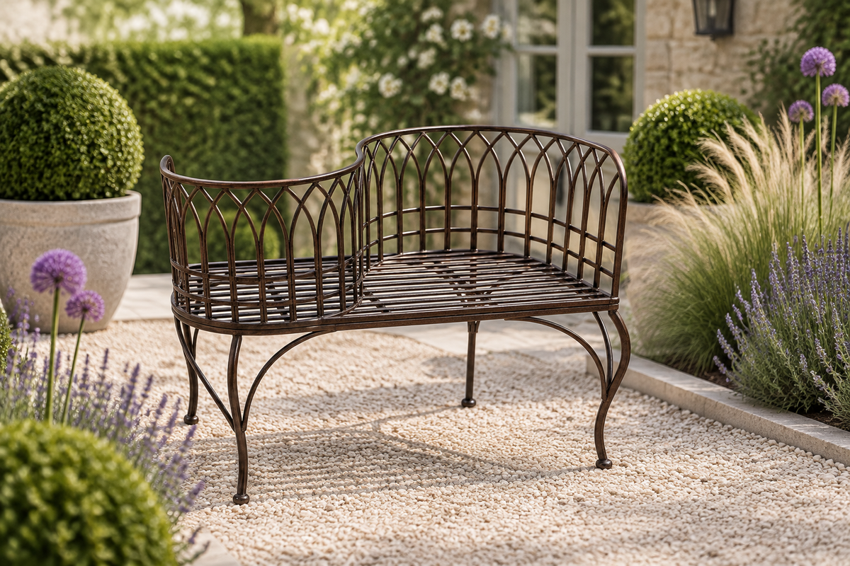 A dark brown wrought iron garden bench with a decorative lattice backrest, set on a gravel path in a landscaped garden with lavender, purple flowers, and green shrubs.