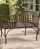 A dark brown metal garden bench with a curved backrest and slatted seat, shown outdoors on a gravel patio in front of a house with hedges and plants.
