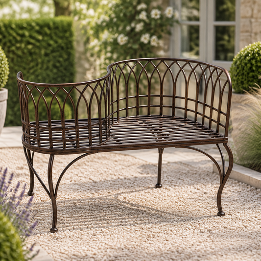 A dark brown metal garden bench with a curved backrest and slatted seat, shown outdoors on a gravel patio in front of a house with hedges and plants.