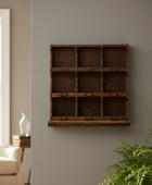 A rustic dark brown wooden wall-mounted organizer with nine compartments, shown hanging on a gray wall in a styled living room setting with a plant and armchair visible.
