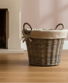Wicker laundry basket with a lid on a wooden floor in front of a fireplace.