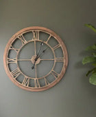 Large round wall clock with Roman numerals on a gray wall next to a plant.