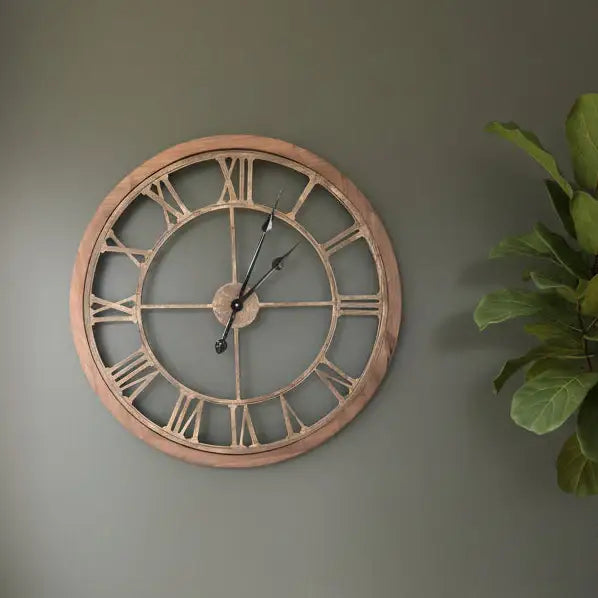 Large round wall clock with Roman numerals on a gray wall next to a plant.
