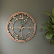 Large round wall clock with Roman numerals on a gray wall next to a plant.