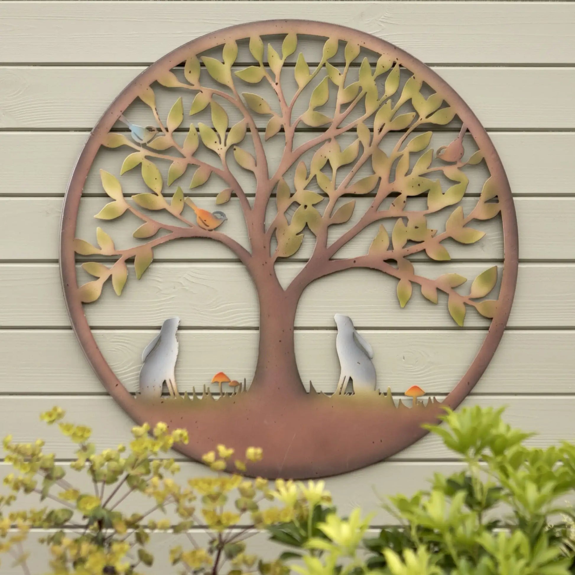 Decorative metal tree wall art on a wooden wall with plants in the foreground.