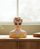 Decorative bust with sunglasses and headband on a wooden surface next to a potted plant and book, with a window in the background.