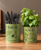 Two olive oil tins styled as home decor on a white marble surface. The left tin is painted sage green and holds gold-colored flatware, while the right tin is a natural olive-green color and contains a green basil plant.