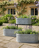 Three metal planters with plants on a stone patio in front of a brick building with flowers.
