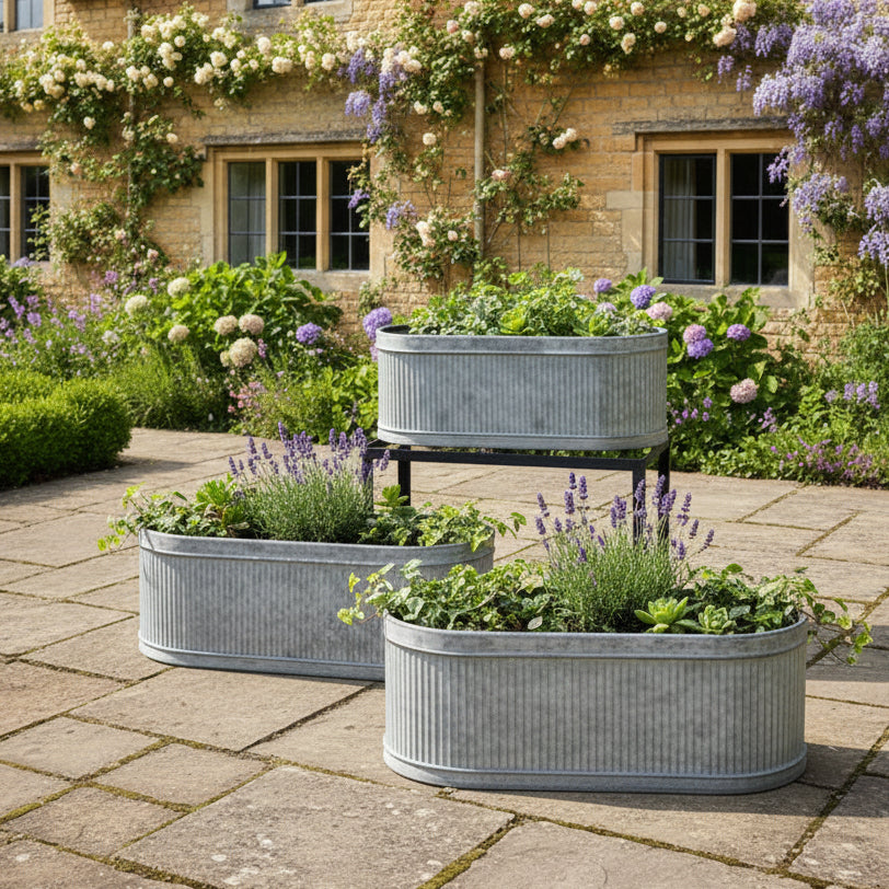 Three metal planters with plants on a stone patio in front of a brick building with flowers.