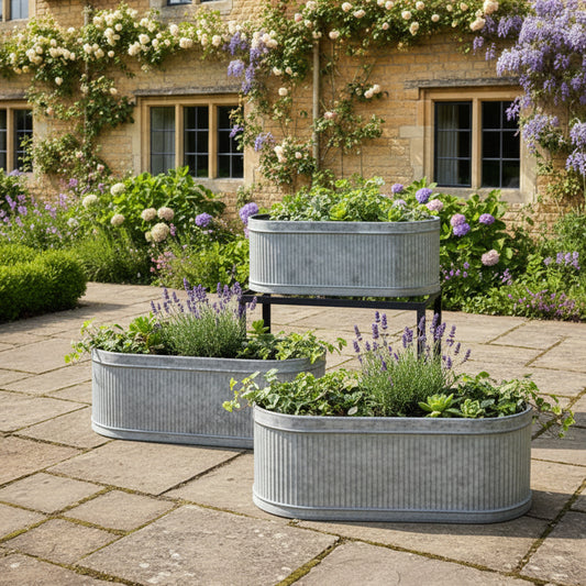 Three metal planters with plants on a stone patio in front of a brick building with flowers.