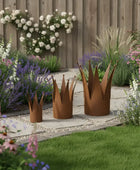 Three metal crown-shaped planters in a garden setting with flowers and a wooden fence.