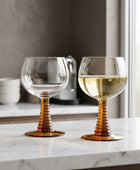 Two clear wine glasses with ribbed amber-brown stems and round bases, placed on a beige-and-white checkered tablecloth. One glass is empty, and the other contains white wine with a dark olive on top.