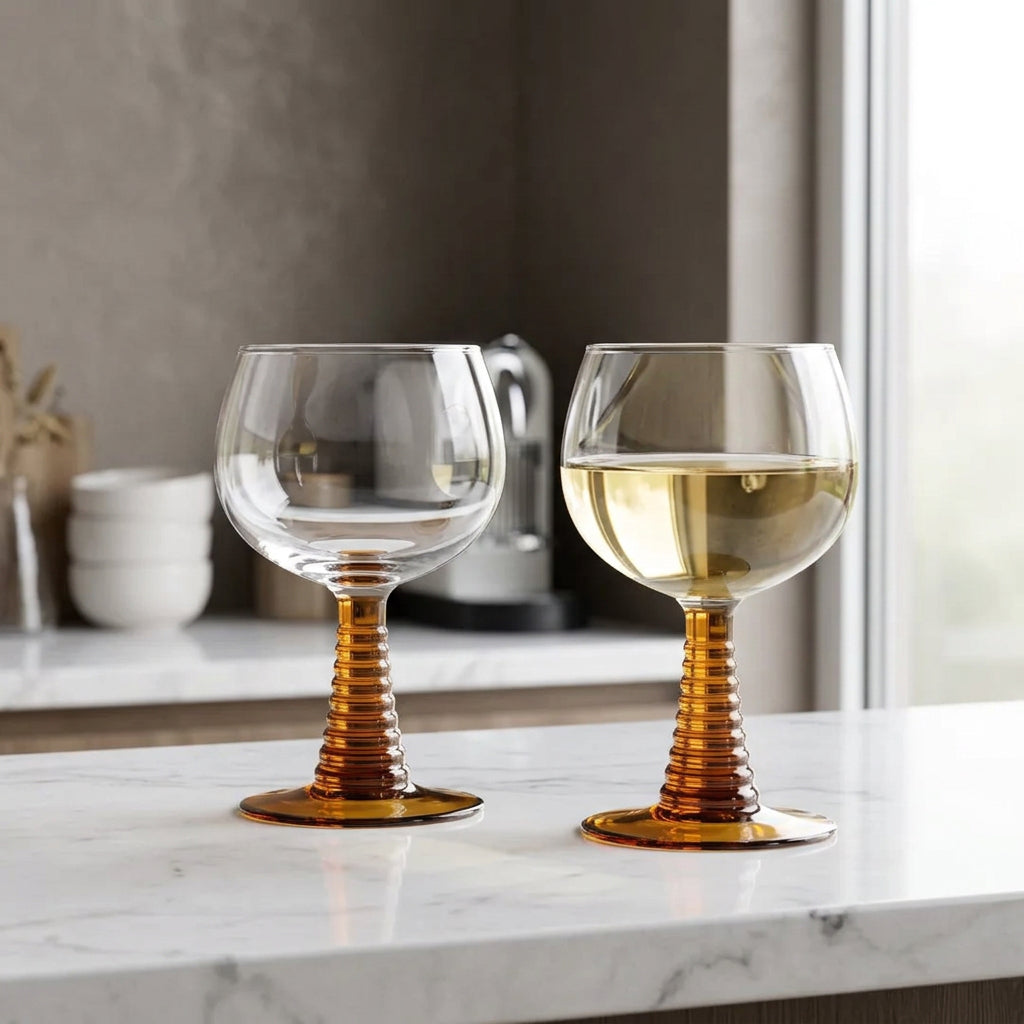 Two clear wine glasses with ribbed amber-brown stems and round bases, placed on a beige-and-white checkered tablecloth. One glass is empty, and the other contains white wine with a dark olive on top.
