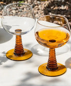 Two stemmed wine glasses with amber-colored ribbed stems and bases, one empty and one partially filled with amber liquid, set on a white table outdoors with gravel in the background.
