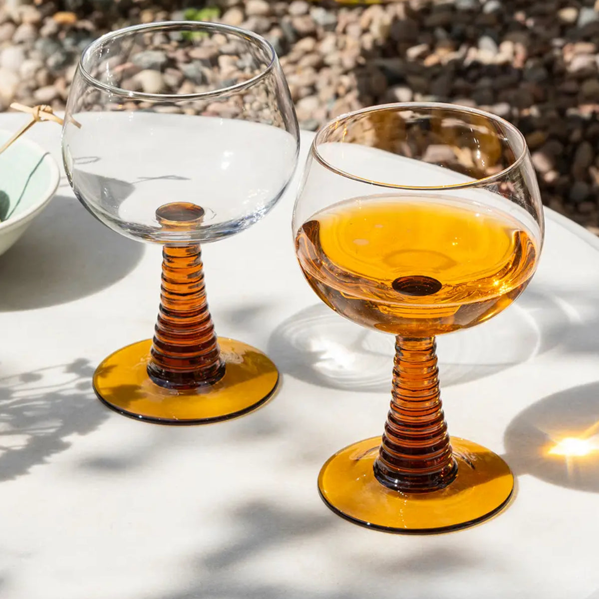 Two stemmed wine glasses with amber-colored ribbed stems and bases, one empty and one partially filled with amber liquid, set on a white table outdoors with gravel in the background.