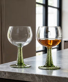 Two stemmed wine glasses with ribbed green stems and clear bowls, placed on a white table outdoors. One glass is empty, and the other contains a light amber drink.