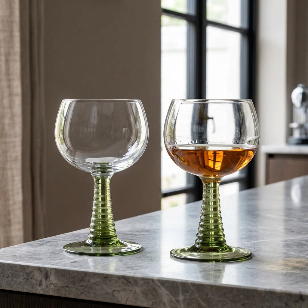 Two stemmed wine glasses with ribbed green stems and clear bowls, placed on a white table outdoors. One glass is empty, and the other contains a light amber drink.