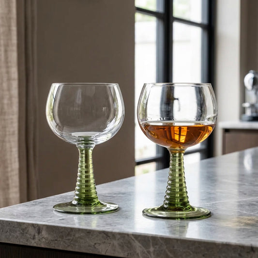 Two stemmed wine glasses with ribbed green stems and clear bowls, placed on a white table outdoors. One glass is empty, and the other contains a light amber drink.