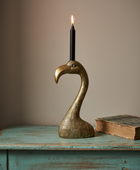 A brass flamingo-shaped candle holder with a black candle inserted, set on a distressed turquoise wooden table against a warm beige background.