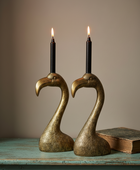 Two brass swan-shaped candle holders with lit black taper candles, displayed on a distressed teal wooden table next to an old book against a neutral beige background.