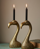 Two brass flamingo-shaped candle holders with black candles, displayed on a rustic teal table next to an old book against a neutral beige background.