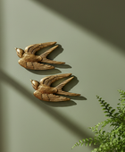 Two decorative swallow wall sculptures in a weathered bronze finish, mounted on a muted sage green wall with soft window light casting shadows and a green plant visible in the lower right corner.