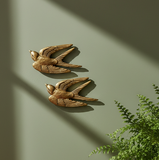 Two decorative swallow wall sculptures in a weathered bronze finish, mounted on a muted sage green wall with soft window light casting shadows and a green plant visible in the lower right corner.