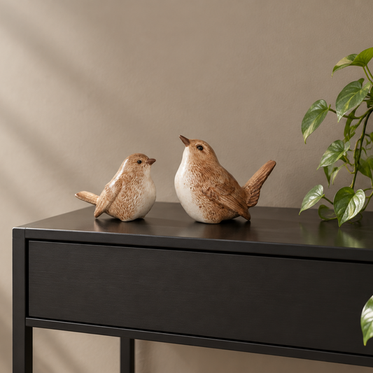 Two ceramic bird figurines, one small cream-and-brown bird and one larger brown-and-white bird, displayed on a black side table against a beige wall with a green plant visible on the right.