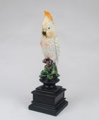 A decorative figurine of a white cockatoo with a bright orange-yellow crest perched on a black pedestal with green leaves, photographed against a neutral light gray background.