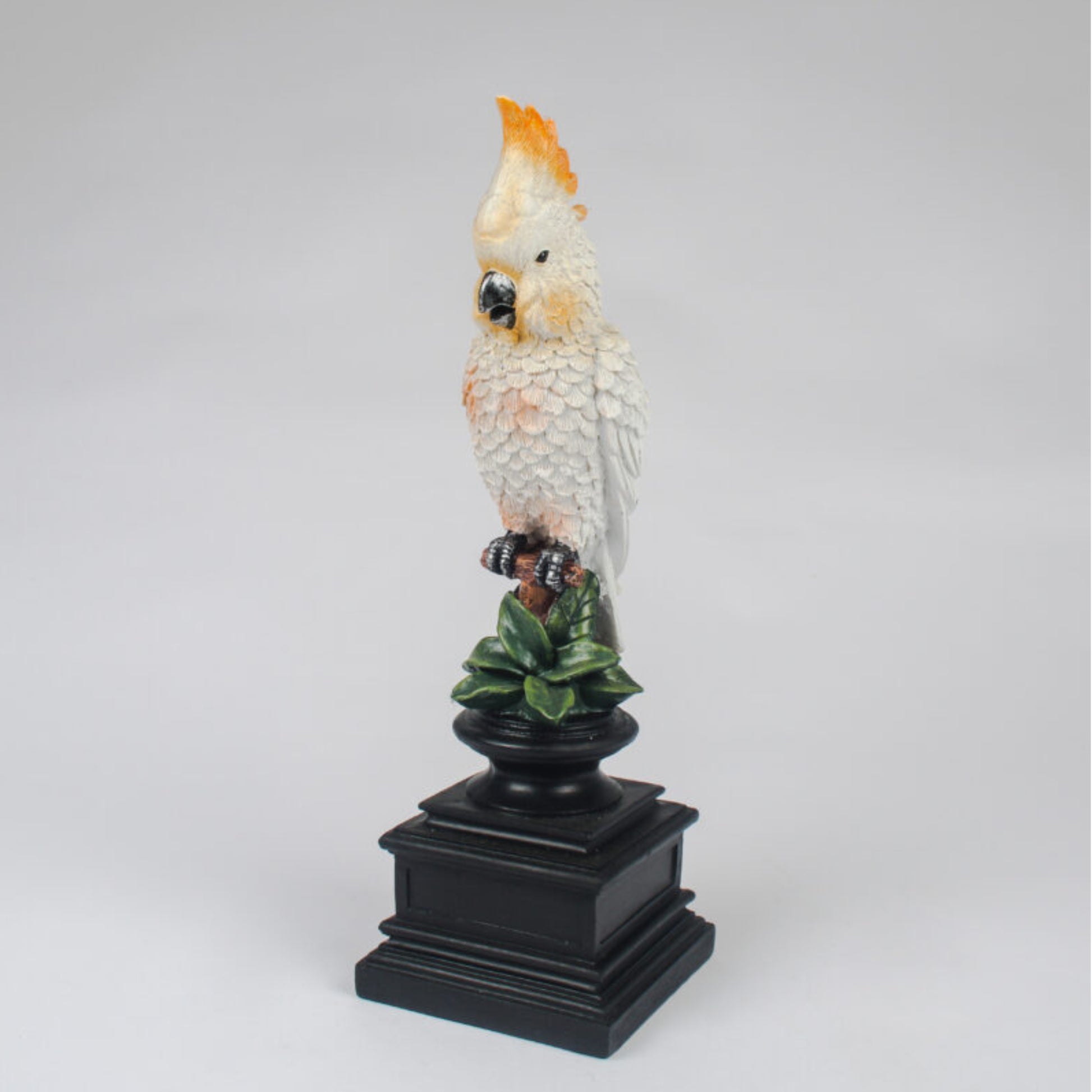 A decorative figurine of a white cockatoo with a bright orange-yellow crest perched on a black pedestal with green leaves, photographed against a neutral light gray background.