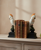 A pair of decorative bookends shaped like white cockatoos with orange crests, perched on black bases and holding a row of vintage-style brown books, displayed on a light wood console table against a beige wall.