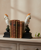 Two decorative cockatoo bookends holding a row of vintage-style books on a light wood cabinet against a beige wall.