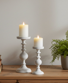 Two white candlesticks with lit candles on a wooden surface, accompanied by a potted plant and books.