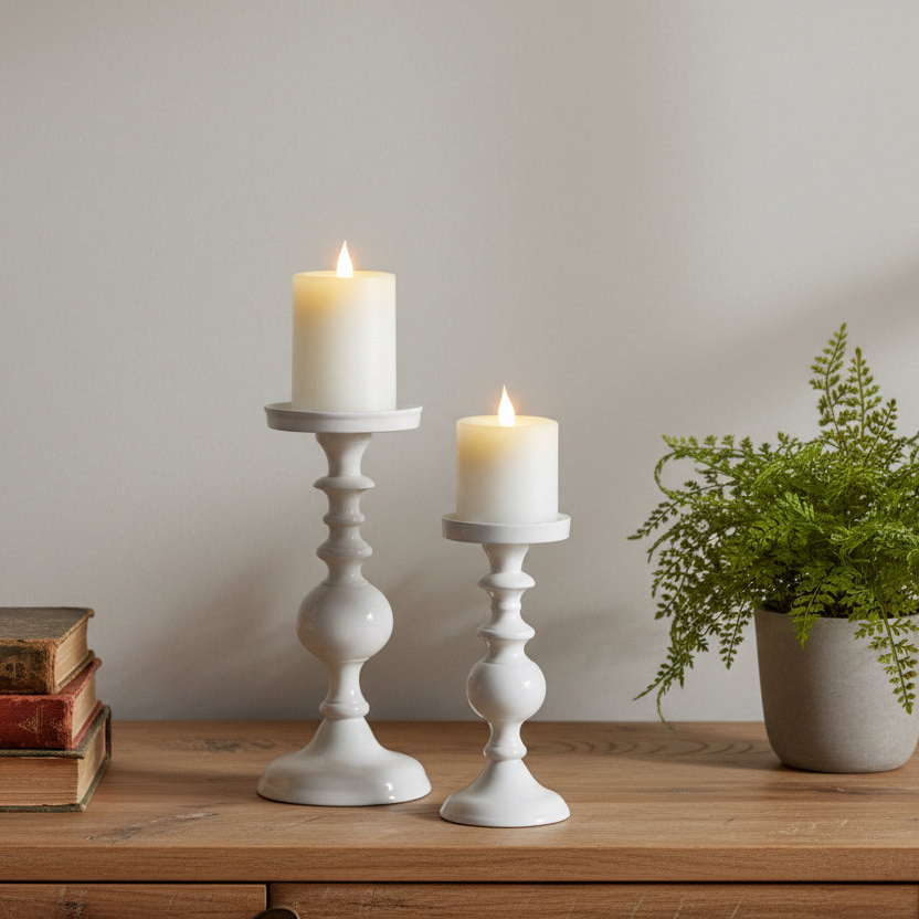 Two white candlesticks with lit candles on a wooden surface, accompanied by a potted plant and books.