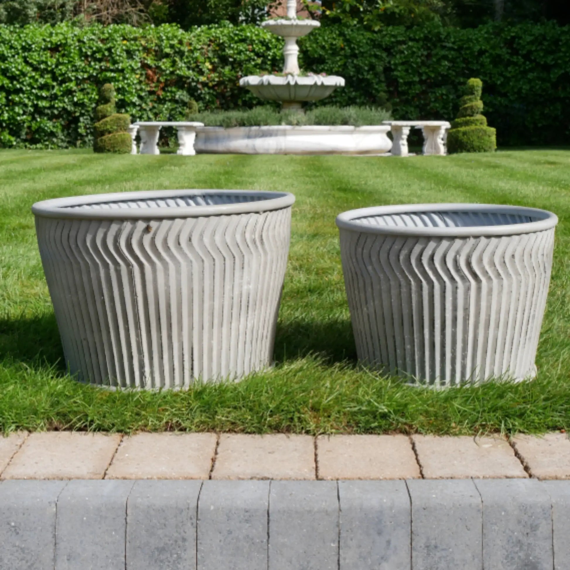 Two concrete planters on grass with a fountain in the background